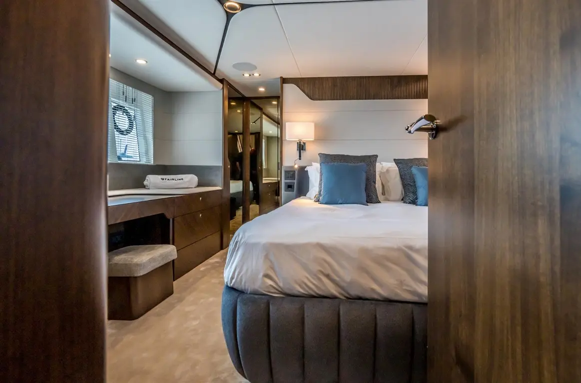 Luxurious yacht cabin featuring a double bed with blue cushions, wooden furniture, and soft lighting; “FAIRLINE” towel visible.